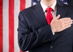 A man in a suit and red tie standing in front of an American flag with his hand on his heart.